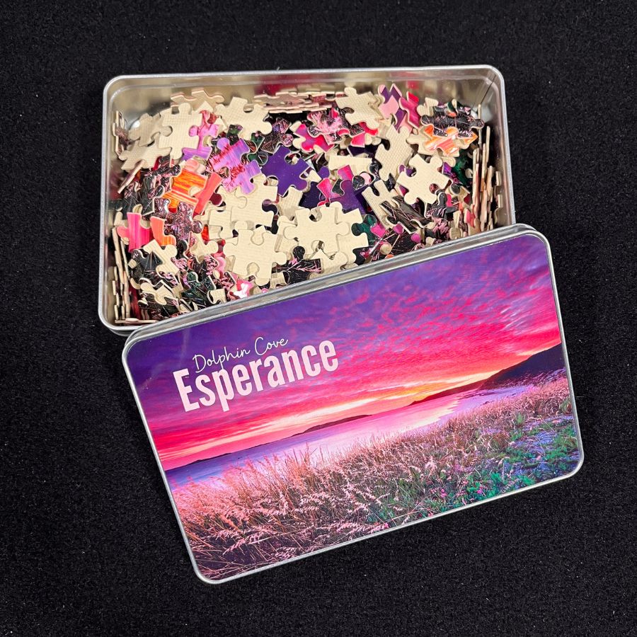 A photo of the 500 piece jigsaw puzzle in pieces inside a storage tin. The jigsaw image is a grassy foreground in front the beach with a red sunset sky.  
