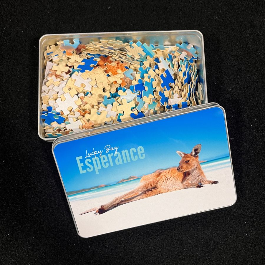 A photo of the 500 piece jigsaw puzzle in pieces inside a storage tin. The jigsaw image is a kangaroo lazing on the beach with a blue sky on a sunny day.  