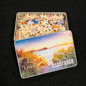 A photo of the 500 piece jigsaw puzzle in pieces inside a storage tin.  it is a sandy path and stair railing with reed grass to the sides and the beach with a sunrise sky, a rock with a hole out in the ocean