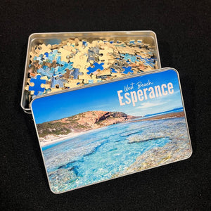 A photo of the 500 piece jigsaw puzzle in pieces inside a storage tin.  The image is a reef pool and a rocky headland with a blue sky on a sunny day.  