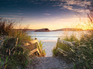 An image of Twilight Cove in Esperance at sunrise showing a golden glow of the sun just out of frame to the right shining onto the stairs and grass leading down a sand track to the beach