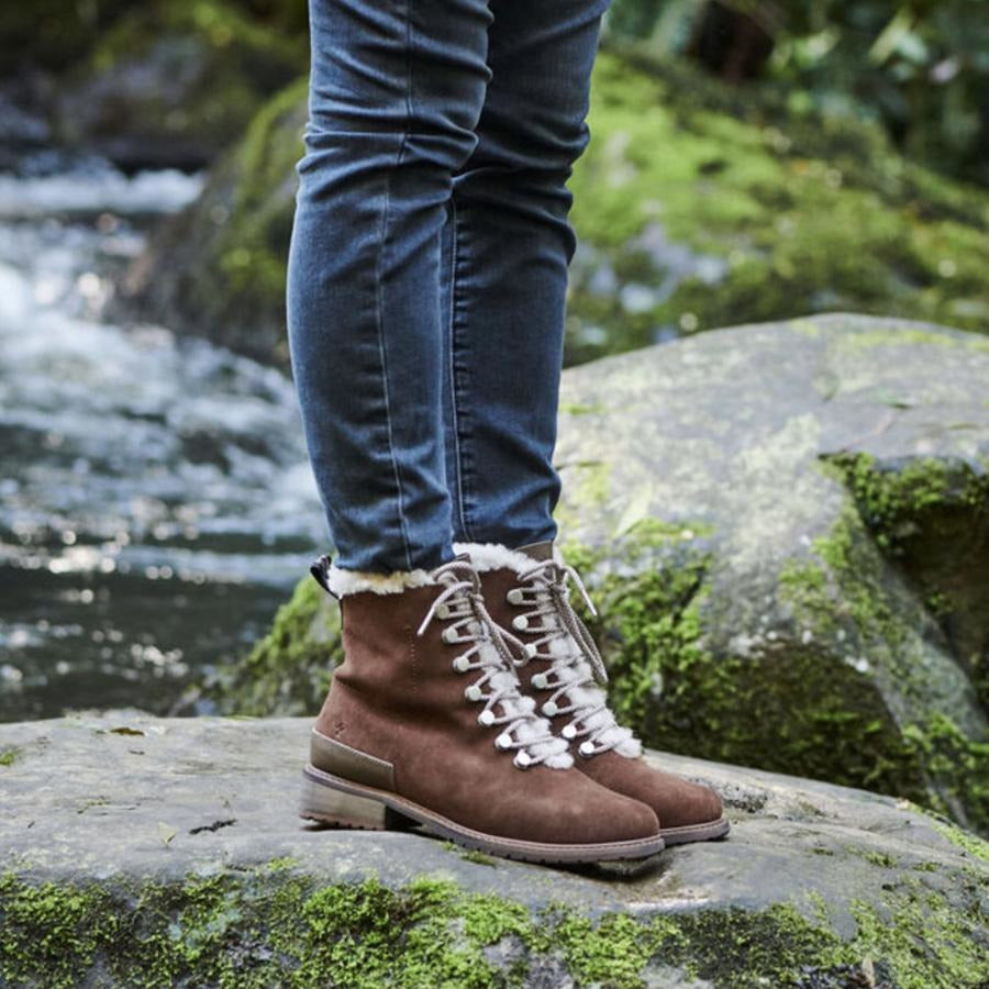 model wearing waterproof fashion wool lined ankle boot oak brown with light brown laces and metal lace hooks standing on a mossy rock with water in background