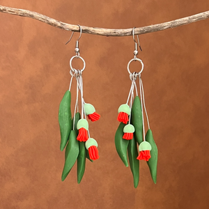 Clay earrings made to look like gum leaves and red blossoms hanging on a stick with a leather background