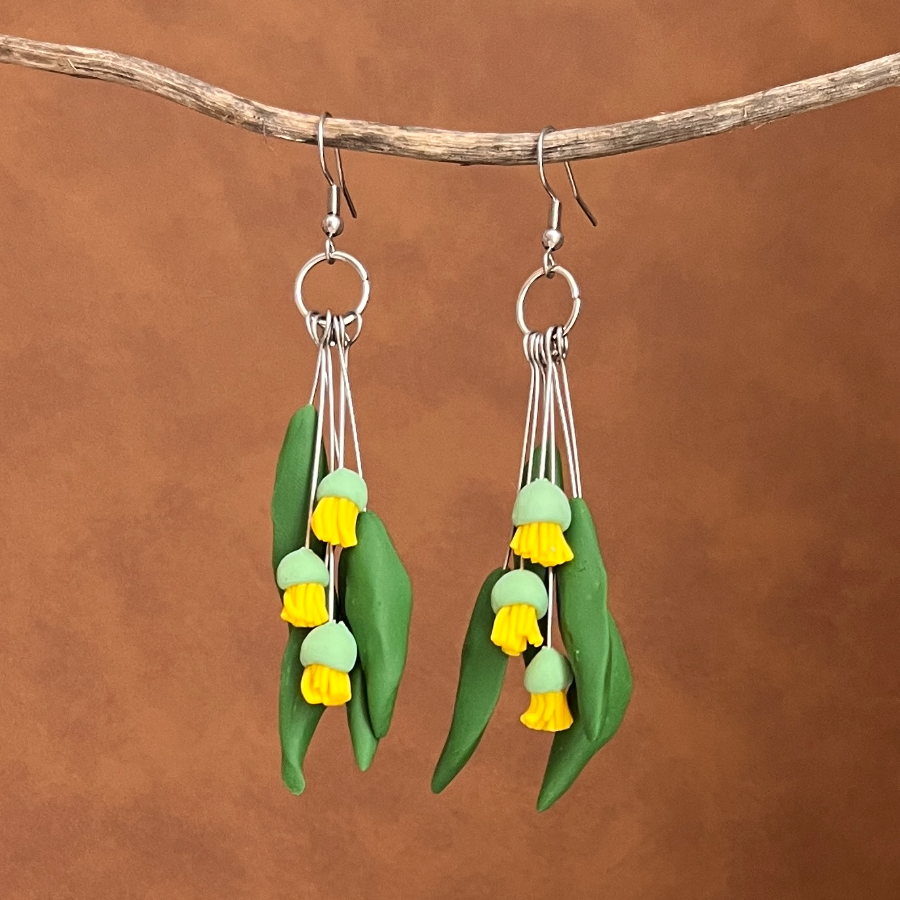Clay earrings made to look like gum leaves and yellow blossoms hanging on a stick with a leather background