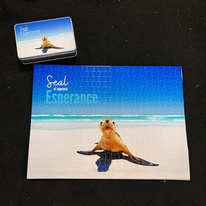 A photo of a 500 piece jigsaw puzzle with a seal on a beach with a blue sky on a sunny day.  Puzzle is complete and the storage tin is next to the puzzle