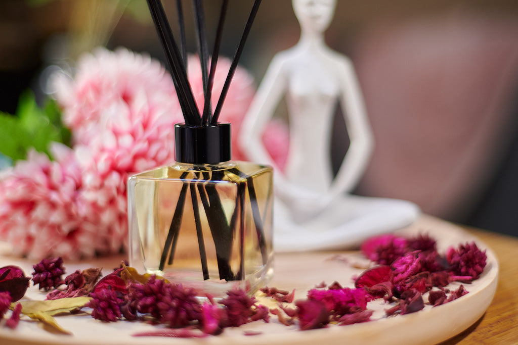 A scented reed diffuser clear glass bottle with a black cap and black reeds displayed on a wooden tray with pink flowers
