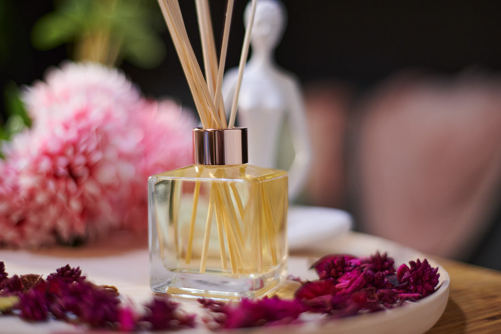 A scented reed diffuser clear glass bottle with a rose gold cap and natural light colour reeds displayed on a wooden tray with pink flowers