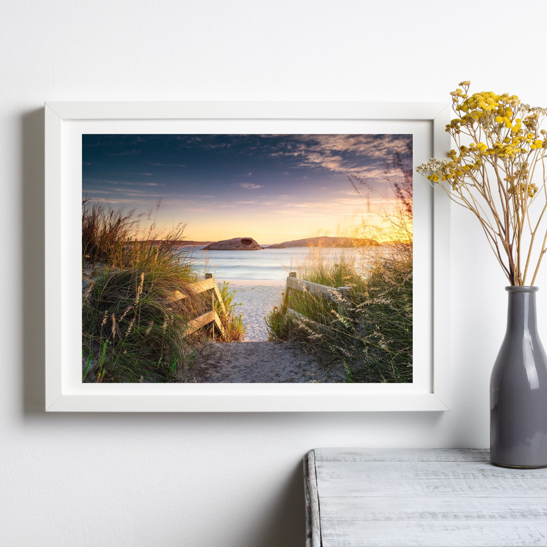 An image of Twilight Cove in Esperance at sunrise showing a golden glow of the sun just out of frame to the right shining onto the stairs and grass leading down a sand track to the beach shown in a white frame hanging on a wall with a vase of yellow flowers to the right