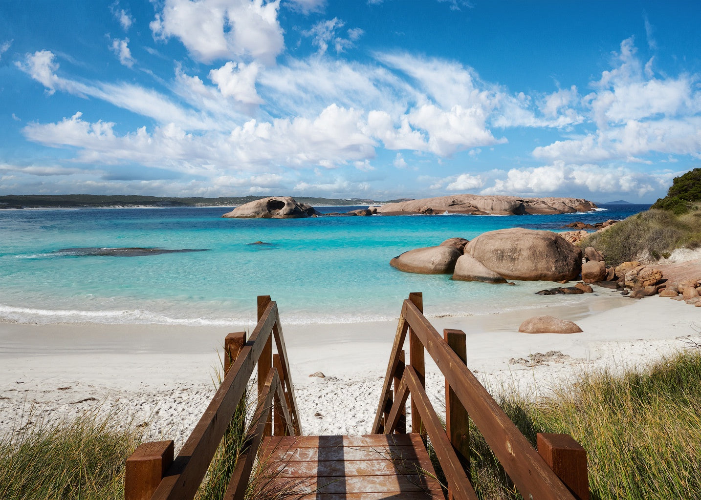 A blue sunny day with whispy clouds, stairs lead down to aqua water and white sand with rocks to the right and a hole in a rock out in the bay