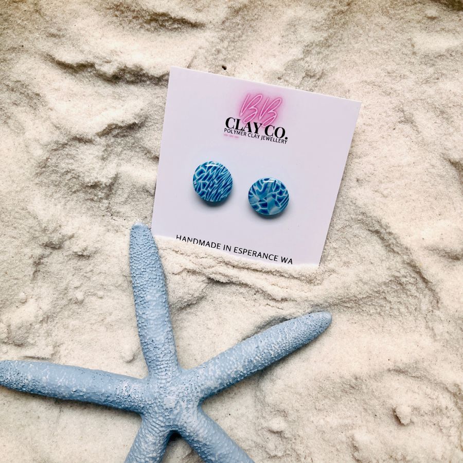 A blue and white pair of earrings that look like the aqua water and white sand displayed on a white backing card, photographed on white sand with a blue starfish