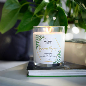 a lit soy wax candle sitting on a book on a bedside table with lush green leaves behind