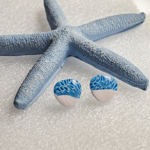 A blue and white pair of earrings that look like the aqua water and white sand, photographed on white sand with a blue starfish