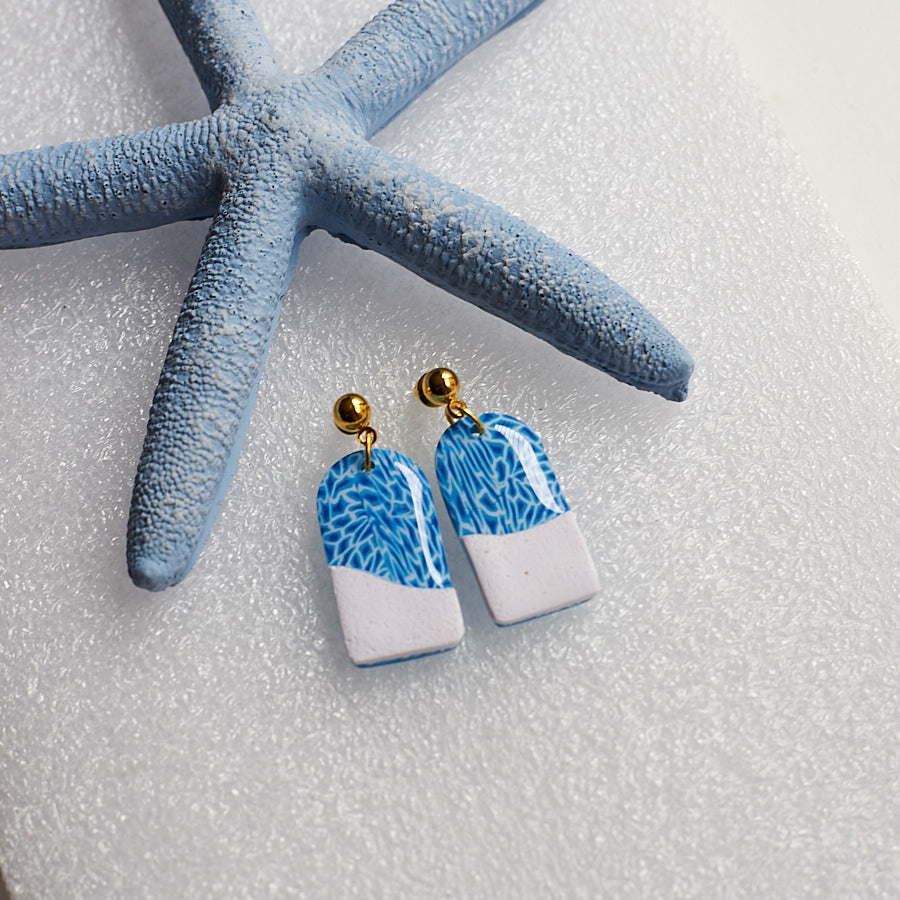 A blue and white pair of earrings that look like the aqua water and white sand, photographed on white sand with a blue starfish