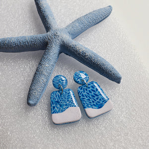 A blue and white pair of earrings that look like the aqua water and white sand, photographed on white sand with a blue starfish